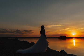 Fototapeta na wymiar Silhouette of bride in white long dress at amazing sunset on sea coast. Sun horizon. Wedding ceremony outdoors on the seashore. Copy space.