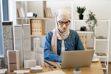 Focused bespectacled muslim female in hijab typing on portable computer at office desk with building models on it. Experienced architect developing urban design using modern gadgets and drawing tools.