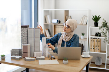 Beautiful arabian female in hijab and glasses arranging building models on office desk during coffee break in creative workplace. Architectural designer analysing details of new project using laptop.