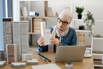 Thoughtful muslim lady in hijab directing building model at laptop camera while working on urban design project in modern workspace. Experienced engineer holding online briefing via video call app.