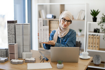 Smiling muslim lady in hijab posing with architectural model in front of laptop while resting from work in creative workplace. Efficient designer planning online presentation of city complex project.
