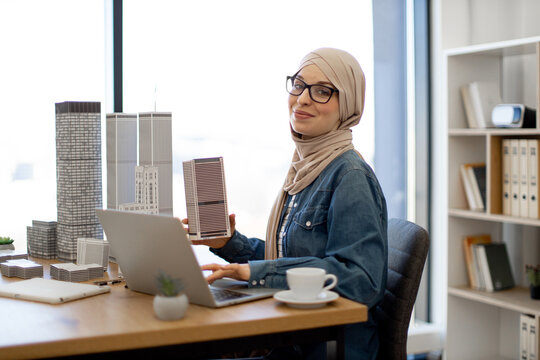 Joyful muslim female wearing hijab and glasses typing on laptop while holding architectural model from office desk indoors. Proficient urban planner developing land use programs of metropolitan area.