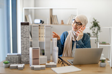 Smiling muslim woman wearing hijab and eyeglasses talking on cell phone while sitting at office desk in creative workspace. Talented architect having business conversation with clients at work.