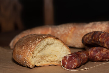 White bread with sesame seeds on it lies on a table. The bread is sliced. In front of it is dried salami, which is also cut. In the background the silhouette of a wooden chair.
