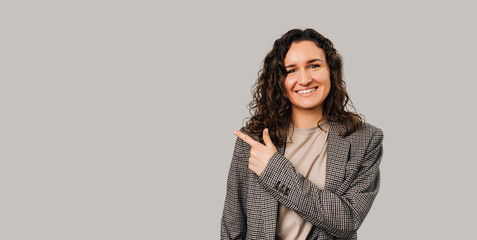 Smart young woman is pointing aside at light grey copy space beside her. Studio portrait.