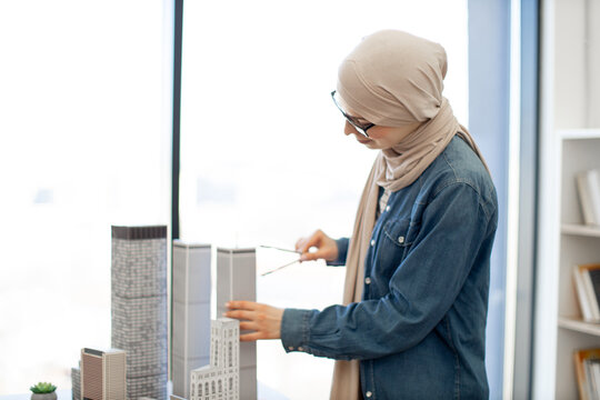 Arabian businesswoman in hijab and spectacles examining building model with compass while standing near writing desk with coffee and gadgets on it. Talanted architect using drawing tools at work.