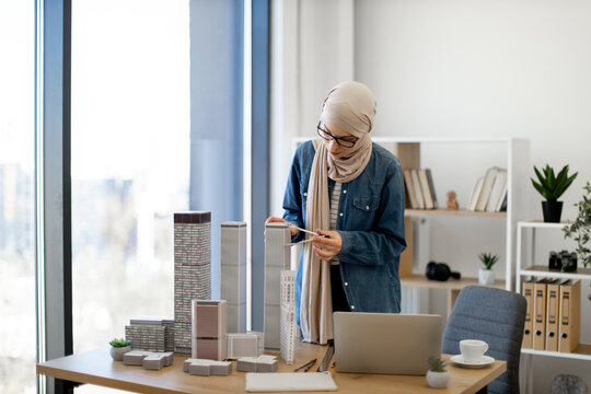 Arabian businesswoman in hijab and spectacles examining building model with compass while standing near writing desk with coffee and gadgets on it. Talanted architect using drawing tools at work.