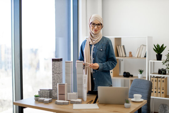 Arabian businesswoman in hijab and spectacles examining building model with compass while standing near writing desk with coffee and gadgets on it. Talanted architect using drawing tools at work. - Powered by Adobe