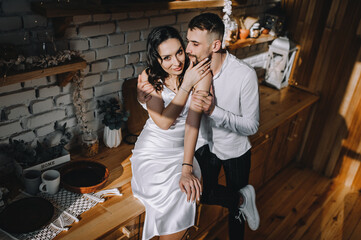 A bearded groom and a beautiful smiling brunette bride are hugging while sitting at a table in a wooden kitchen in a cafe interior in sunlight. Wedding photography of happy newlyweds, portrait.