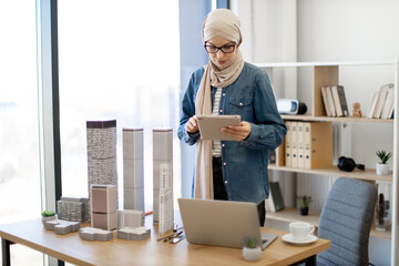 Smiling arabian lady in beige hijab working on digital tablet with open laptop placed near architectural models on office desk. Design professional developing construction project using technologies.