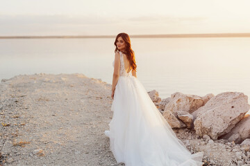 Fototapeta na wymiar Happy young bride in a white long dress walks on the shore of the azure sea at sunset. Outdoor wedding ceremony on the seashore.