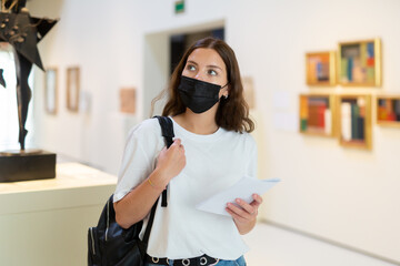 Attentive visitor girl in a protective mask, who is in the museum during the pandemic with an...
