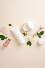 Elevate your self-care routine with top view vertical cosmetology concept featuring pump bottles of face roller serum and cream, massage rollers, and green leaves on a serene pastel beige backdrop