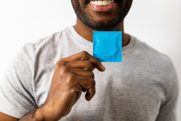 An unrecognizable dark-skinned man is holding a condom while smiling in a casual gray shirt,...