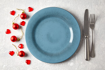 Empty blue ceramic plate with cutlery surrounded by red christmas balls and hearts on a light concrete table