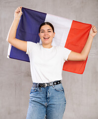 Glad young woman in casual clothes posing and smiling, standing with flag of France against gray...