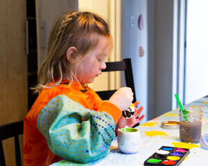 Selective focus medium portrait of fair little girl in art waterproof smock painting an egg for a...