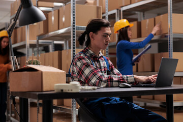 Young asian warehouse supervisor using laptop in storage room. Storehouse worker supervising...