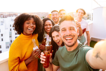 group of young people taking a selfie on terrace while having a party. They are happy and toasting with beer bottles. 