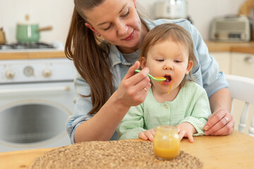 Happy family at home. Mother feeding her baby girl from spoon in kitchen. Little toddler child with messy funny face eats healthy food at home. Young woman mom giving food to kid daughter.