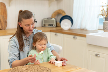Happy family at home. Mother feeding her baby girl from spoon in kitchen. Little toddler child with messy funny face eats healthy food at home. Young woman mom giving food to kid daughter.