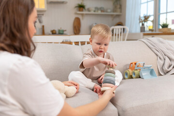 Happy family at home. Mother and baby boy playing with toys in couch at home indoors. Little toddler child and babysitter nanny having fun together. Young woman mom kid son rest in living room. - obrazy, fototapety, plakaty