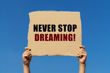Never stop dreaming text on box paper held by 2 hands with isolated blue sky background. This message board can be used as business concept about success.