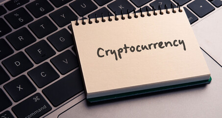 There is note book with the word Cryptocurrency on a laptop. It is an eye-catching image.