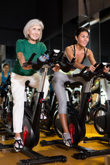 Fototapeta na wymiar Young adult and elderly women training together during stationary bike workout in fitness center