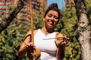 mujer morena tocando berimbau instrumento de percusión de capoeira, en un parque con un día...