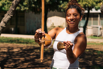 mujer morena tocando berimbau instrumento de percusión de capoeira, en un parque con un día...