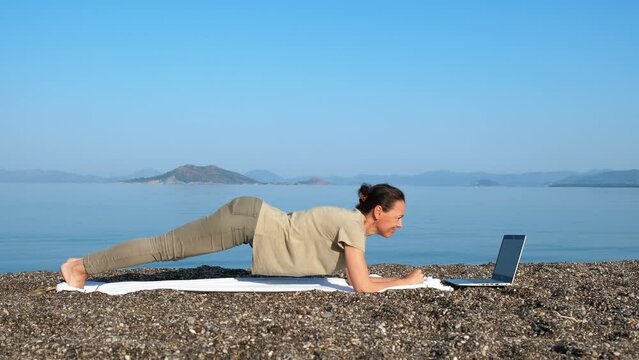 Four limbed staff pose on shore. A happy sporty woman practicing four limbed staff pose by the laptop on the luxury beach. A concept of sporty activity during summer traveling.