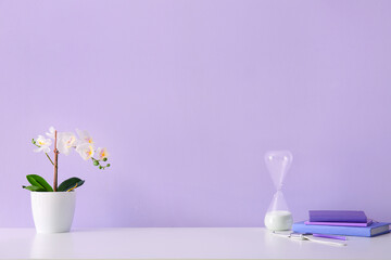 Workplace with flower, hourglass, smartwatch and books near lilac wall