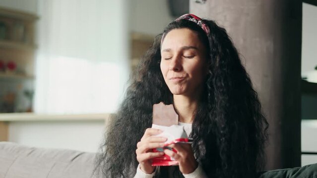 Portrait of young hispanic woman eating chocolate and having toothache sitting on sofa at home Sad female feeling pain in tooth because caries gum disease or sensitive teeth indoors