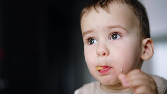 Cute baby boy stuck a piece of cookie into mouth. Toddler tries hard to chew the biscuit as he’s got banana in hand to eat next. Close up. Blurred backdrop.