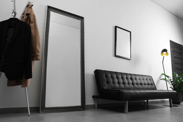 Coat rack, stylish leather sofa and large mirror near white wall in hallway. Interior design