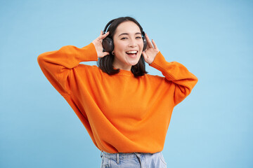 Happy Chinese woman in headphones, listens music, enjoys favourite song in her playlist, stands over blue background