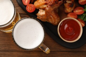 Mugs with beer, delicious baked chicken wings and sauce on wooden table, flat lay