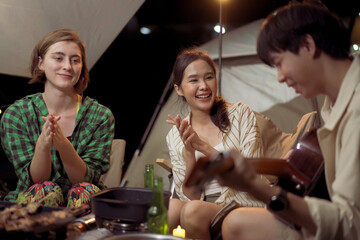 Group of diverse ethnicity young people enjoy eating and grilling bar-b-q together while camping in forest at night. Happy friends enjoy drinking a beer and playing   a guitar between camping.