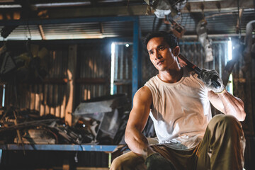 Young and handsome muscular and strop scrap material man in sleeveless shirt sitting on material in junkyard while looking away while taking rest and break from work during in warehouse