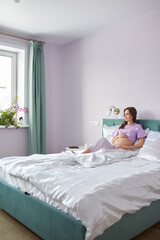 A pregnant woman sits in bed of her domestic bedroom and looking oout the window. Female enjoying her pregnancy state.