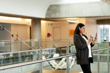 Asian mature woman using smartphone, standing by stairs in modern office interior. Technology People