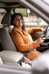 Fototapeta na wymiar Side portrait of confident mature Indian businesswoman in casual clothes, sitting in modern car on driver's seat, smiling looking at camera. Business, successful people, transportation concept 