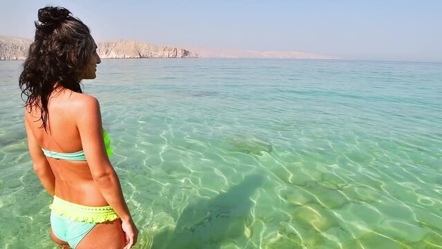 Caucasian brunette tourist woman in bikini stand on white sandy beach crystal clear turquoise water alone on vacation in persian gulf Mirellas island. Musandam.Oman