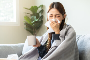 Sick, hurt or pain asian young woman have a fever, flu sore throat, drinking a mug of warm water, use tissues paper sneezing nose, runny while sitting rest on sofa at home. Health care on virus person