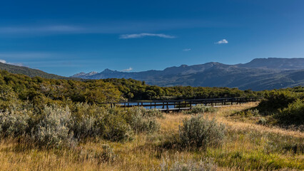 A wooden pedestrian path runs in the valley next to the lake. Bushes and yellowed grass grow in the meadow. Picturesque mountains against the blue sky. Argentina. Tierra del Fuego National Park.