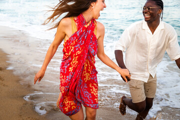 Young multiracial happy couple running along seaside during vacation travel holding hands.