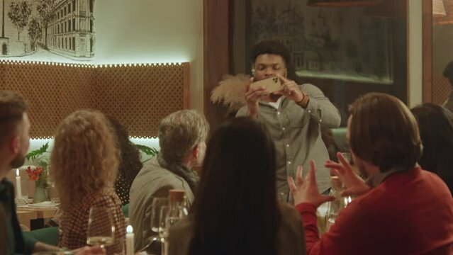 African American man takes photo and selfie with friends in cozy cafe using phone. Group of diverse people spend evening together, hangout on birthday party in restaurant. Concept of public eating.