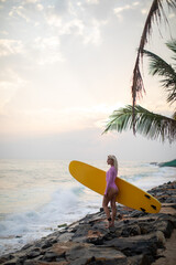 Female surfer in a pink swimsuit and with a yellow surfboard on a rocky beach.