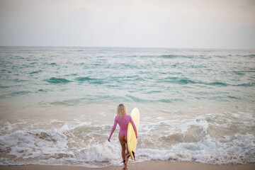 A woman surfer in a pink swimsuit and with a yellow surfboard enters the ocean.